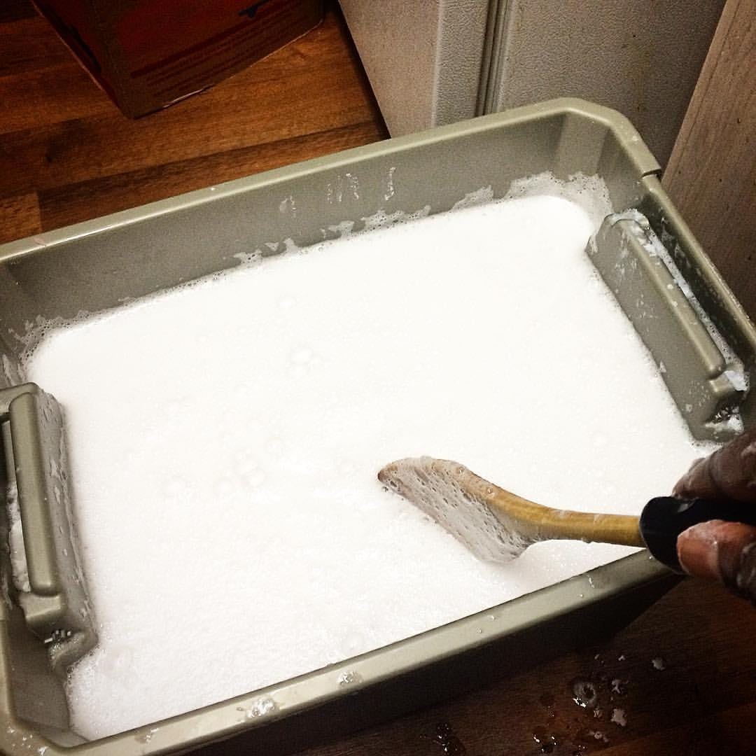 A big ole batch of Homemade Laundry Detergent in a grey 7.5 gallon bin. The thumb of a Black persona's hand is holding a wooden spoon immersed in the suds.